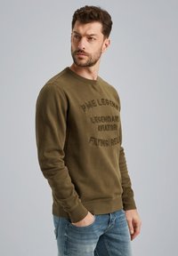 Oliwkowy sweter z miękkiego materiału. Wyróżnia się wytłoczonym tekstem na przodzie, prążkowanymi mankietami i dołem, w luźnym kroju.