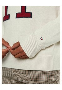 Dzianinowy kremowy sweter z czerwonymi i granatowymi cyframi. Posiada ściągacze przy mankietach i u dołu, z małym akcentem logo blisko rękawa.