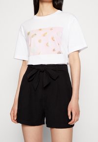 Weißes T-Shirt mit einem grafischen Motiv im pinken Muschelmuster, kombiniert mit hochtaillierten schwarzen Shorts mit einer vorderen Schleifenverzierung und glatter Textur.