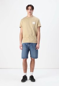 Beige cotton t-shirt with a small, white square patch. Paired with blue denim shorts. Black sneakers and white socks. Neutral background.