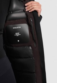 Capa interna acolchada negra de una chaqueta, con una etiqueta de tela que muestra el nombre de la marca "KHUJOTM" y el año de establecimiento, textura suave y diseño elegante.