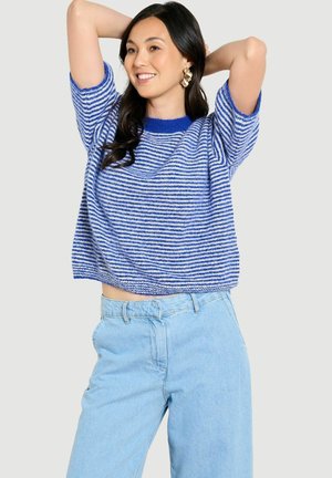Frau lächelt mit erhobenen Armen hinter dem Kopf, trägt einen blau-weiß gestreiften Kurzarmpullover und hellblaue High-Waist-Jeans.