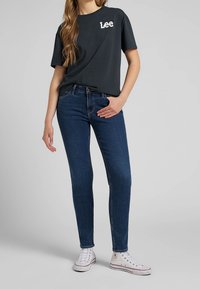 Dark gray cotton t-shirt with white "Lee" logo, paired with blue denim skinny jeans and white canvas sneakers. Simple design, casual fit.