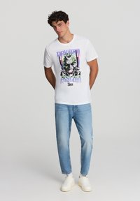 Biała bawełniana koszulka z grafika przedstawiająca postać z zielonymi włosami, napis "Arkham Asylum", z kolorowymi akcentami i okrągłym dekoltem.