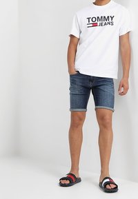 Biała bawełniana koszulka z logo "TOMMY JEANS", niebieskie dżinsowe szorty z podwiniętymi brzegami oraz czarne klapki z czerwonymi i białymi paskami.