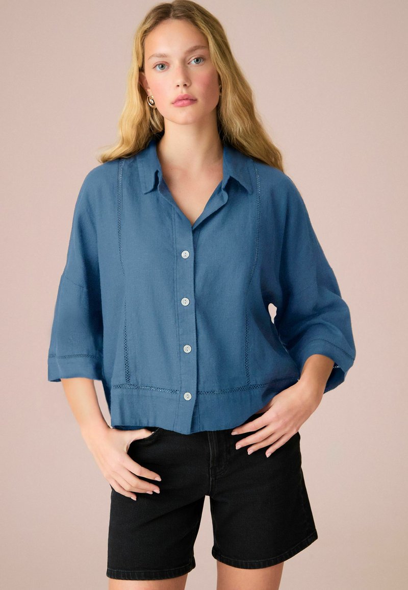 Chemise bleue à manches courtes avec un col, des coutures décoratives et des boutons clairs ; portée avec un short noir.