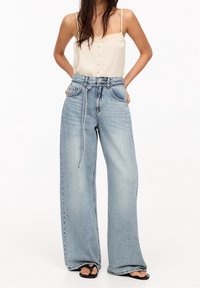 Mujer vestida con pantalones vaqueros de pierna ancha de color azul claro con un cinturón de cordón, una blusa sin mangas beige y sandalias de dedo negras, de pie con las manos en los bolsillos.