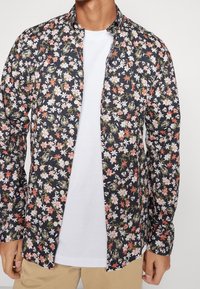 Chemise à boutons florale en bleu marine foncé avec des motifs de fleurs roses, blanches et vertes. Tissu léger, manches longues et col classique.