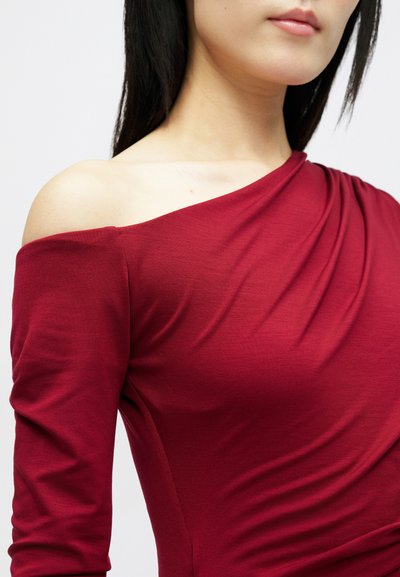 Mulher a usar uma blusa vermelha com decote apenas num dos ombros e mangas até ao cotovelo, mostrando um ombro exposto e parte do pescoço e dos lábios.