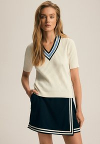 Beżowy bezrękawnik dzianinowy o prążkowanej fakturze i dekolcie w kształcie litery V, w połączeniu z granatową spódnicą z białymi paskami wzdłuż hemu.