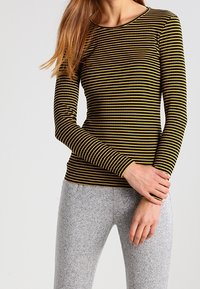 Långärmad topp i svart och gula ränder, med rundad halslinje och en kroppsnära passform. Bärs med grå, strukturerade leggings.