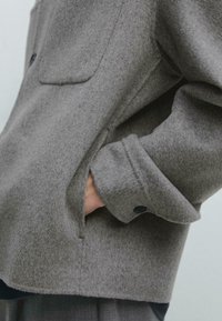 Grå uldjakke med en struktureret overflade, der har en brystlomme, knapmanchetter og sidelommer. Designet har en afslappet pasform.