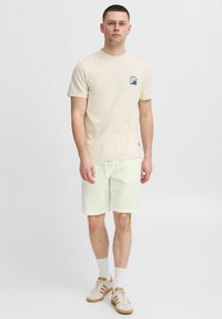 Beige pamut póló, kicsi kék és fehér grafikával, világos lennadrággal és fehér sportcipővel, arany csíkokkal párosítva.