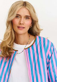 pikowana kurtka bomberka w niebiesko-różowe paski z brązowymi wykończeniami, z zamkiem błyskawicznym z przodu i białym T-shirtem z okrągłym dekoltem pod spodem.