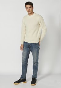 Crème-kleurige gebreide trui met een textuurpatroon, gecombineerd met blauwe denim jeans en zwarte sneakers met een gumrubberen zool.
