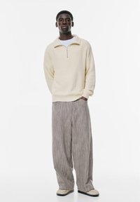 Beige gerippter Pulli mit einem Reißverschlusskragen, kombiniert mit hellgrauen gestreiften weit geschnittenen Hosen und beigen Slippern. Einfaches, entspanntes Design.
