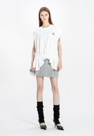 Joven mujer de pie con top blanco sin mangas de gran tamaño con dobladillo fruncido, falda gris en capas, calentadores de piernas negros fruncidos y tacones negros.