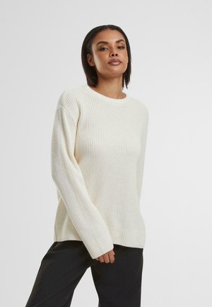 Cream-colored knit sweater with a ribbed texture, crew neckline, and long sleeves. Notable for its relaxed fit and soft fabric.