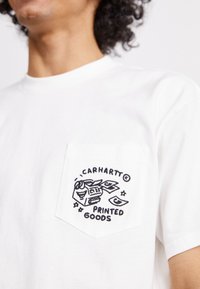 Bijela pamučna majica s crnim izvezenim džepom sa natpisom "Carhartt Printed Goods" i maštovitim grafičkim motivima.