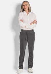 Vrouw met lang grijs haar staat met gekruiste armen, draagt een witte blouse, grijze drawstring broek en witte sneakers op een witte achtergrond.