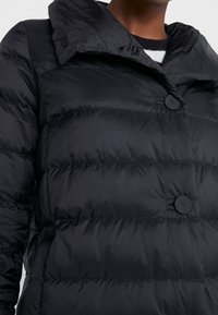 Sort puffjakke med stor krave og to synlige runde knapper over en sort-hvid stribet sweater.