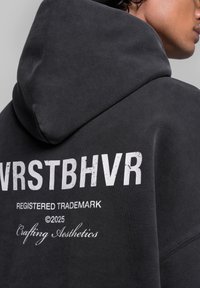Felpa grigia con un grande logo bianco "VRSTBHV" e testo sottostante. Presenta un cappuccio con lacci e una consistenza morbida. Dettaglio delle cuciture visibile.