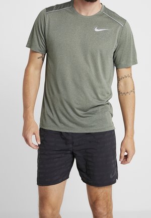 Muž v olivově zeleném sportovním tričku s krátkým rukávem značky Nike a černých kratších Nike šortkách s potiskem "Just Do It", stojící před jednobarevným světlým pozadím.