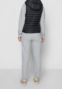 Chaleco acolchado negro con capucha sobre una camiseta de manga larga gris claro y pantalones de chándal a juego, combinados con zapatos beige. Tejido suave y texturizado.