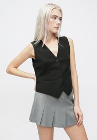 Μαύρο αμάνικο γιλέκο με V-λαιμόκοψη, πέντε κουμπιά και δύο μπροστινές τσέπες, συνδυασμένο με ανοι�χτό γκρι μίνι φούστα με πιέτες.