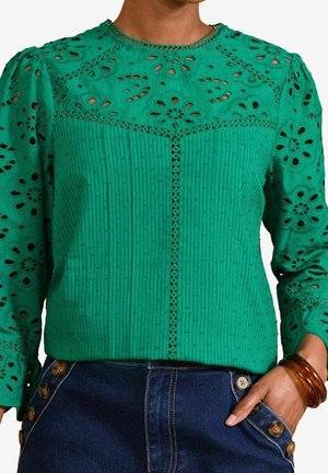 Femme portant un chemisier en dentelle ajourée verte à manches longues et un jean bleu taille haute boutonné, la main dans la poche.