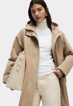 Manteau de pluie beige avec une capuche, fermeture éclair et cordon de serrage réglable. Associé à un col roulé blanc à côtes et à un pantalon beige, tenant un sac assorti.