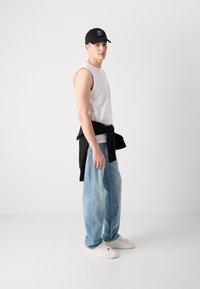 Camiseta sin mangas blanca, jeans de mezclilla sueltos azul claro, sudadera negra atada a la cintura, zapatillas blancas, gorra negra con texto.