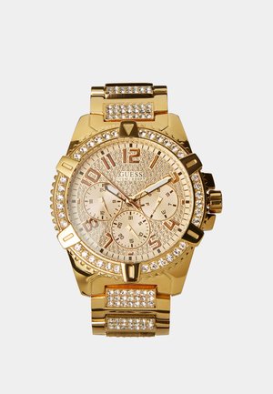 Guess polshorloge in goudkleur met kristallen accenten, drie subwijzers, grote cijfers en een schakeltekenarmband met extra kristallen.