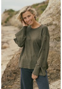 Pull vert olive avec un col rond, des manches larges et des accents de boutons le long des bras. Confectionné dans un tissu tricoté doux, il présente une coupe décontractée.