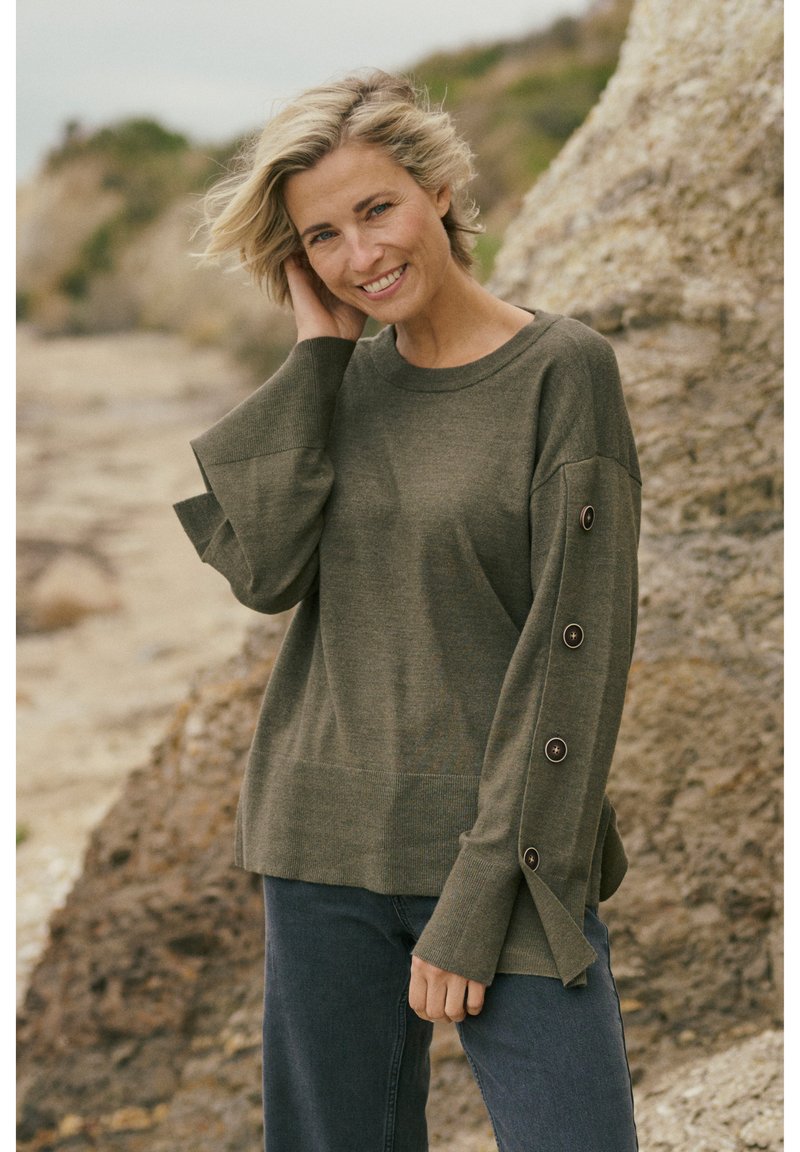 Pull vert olive avec un col rond, des manches larges et des accents de boutons le long des bras. Confectionné dans un tissu tricoté doux, il présente une coupe décontractée.