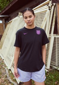 Czarna koszulka Nike z fioletowym logo i herbem, w zestawie z białymi i granatowymi spodniami w paski. W tle drewniane konstrukcje i zieleń.