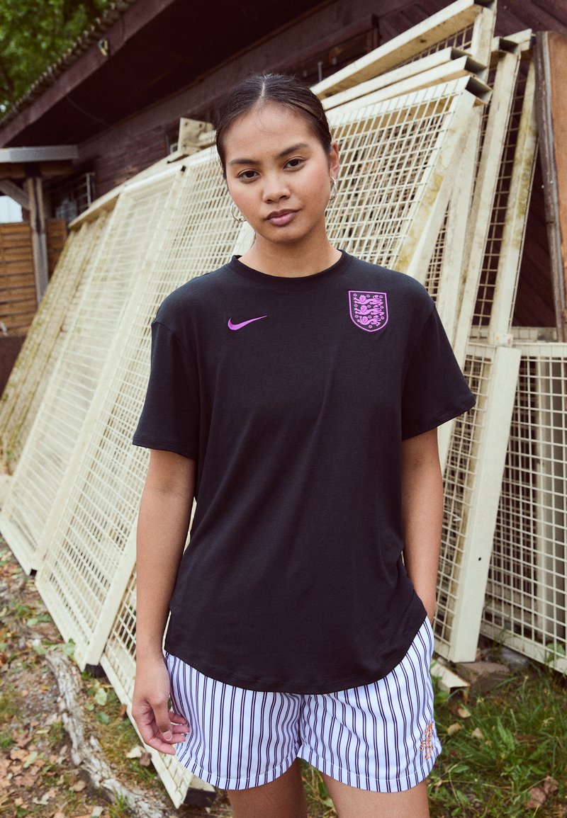 Melnā Kurzā Nike T-kreklā ar purpursarkanām emblēmām; savienots ar svītrainām šortiem. Fons ietver koka konstrukcijas un zaļumu.