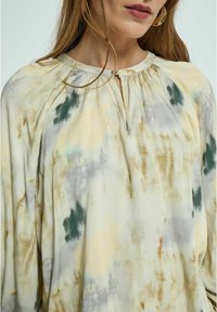 Ľahká blúzka s tie-dye vzorom v pastelových odtieňoch žltej, zelenej a sivej. Má okrúhly výstrih s uzáverom v tvare kľúčovej dierky a skladané ramená.