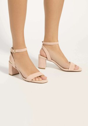 Voeten met lichtroze sandalen met blokhak, enkelbandjes en gespen op een effen lichte achtergrond.