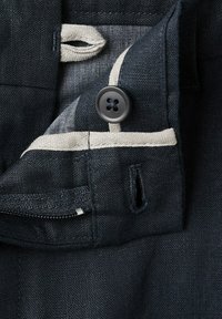 Primo piano di tessuto scuro con un bottone nero e dettagli di rifinitura beige, che mostra un occhiello e una cerniera su un indumento.