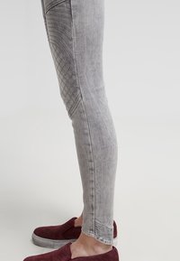 Vaqueros ajustados grises con paneles laterales texturizados con un patrón de diamante y costuras detalladas, combinados con zapatos tipo slip-on en color granate.