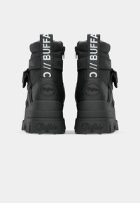 Black ankle boots with a textured finish, featuring a logo strap, zip closure, and rugged soles for traction. Reinforced heel design.
