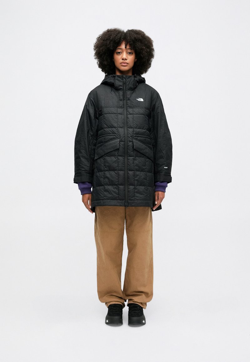 Czarna ocieplana kurtka z kapturem, z pikowanym wzorem, kieszeniami z przodu i logo The North Face na klatce piersiowej. Noszona z beżowymi spodniami.