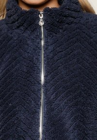 Marineblaue Fleecejacke mit hohem Kragen, ausgestattet mit einem Zickzack-Muster und einem Frontreißverschluss mit Metallbeschlägen.