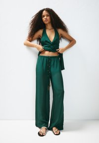 Zelený saténový halter top s uzlovým detailem, doplněný širokými kalhotami s pozlacenými knoflíky a stahovací šňůrkou v pase. Černé sandály.