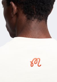 Vue arrière d'une personne aux cheveux courts et bouclés portant une chemise blanche avec un petit logo orange brodé près du col.