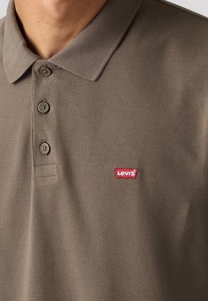 Man draagt een bruin poloshirt met drie knopen en een klein rood Levi's-logo op de linkerborst.