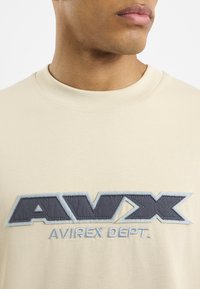 Kremowa koszulka z krótkim rękawem i okrągłym dekoltem, z wyraźnym granatowym haftowanym logo, które brzmi "AVIREX DEPT." w odważnym stylu.