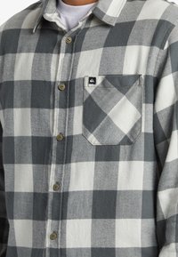 Camicia a quadri in flanella grigia e bianca, con chiusura a bottoni sul davanti, una tasca sul petto e un piccolo etichetta con il logo sulla tasca.