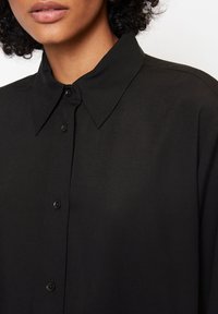Schwarzes Hemd mit Knopfleiste, ausgestattet mit einem klassischen Kragen und glatter Textur. Enthält schwarze Knöpfe und einen lockeren Schnitt.
